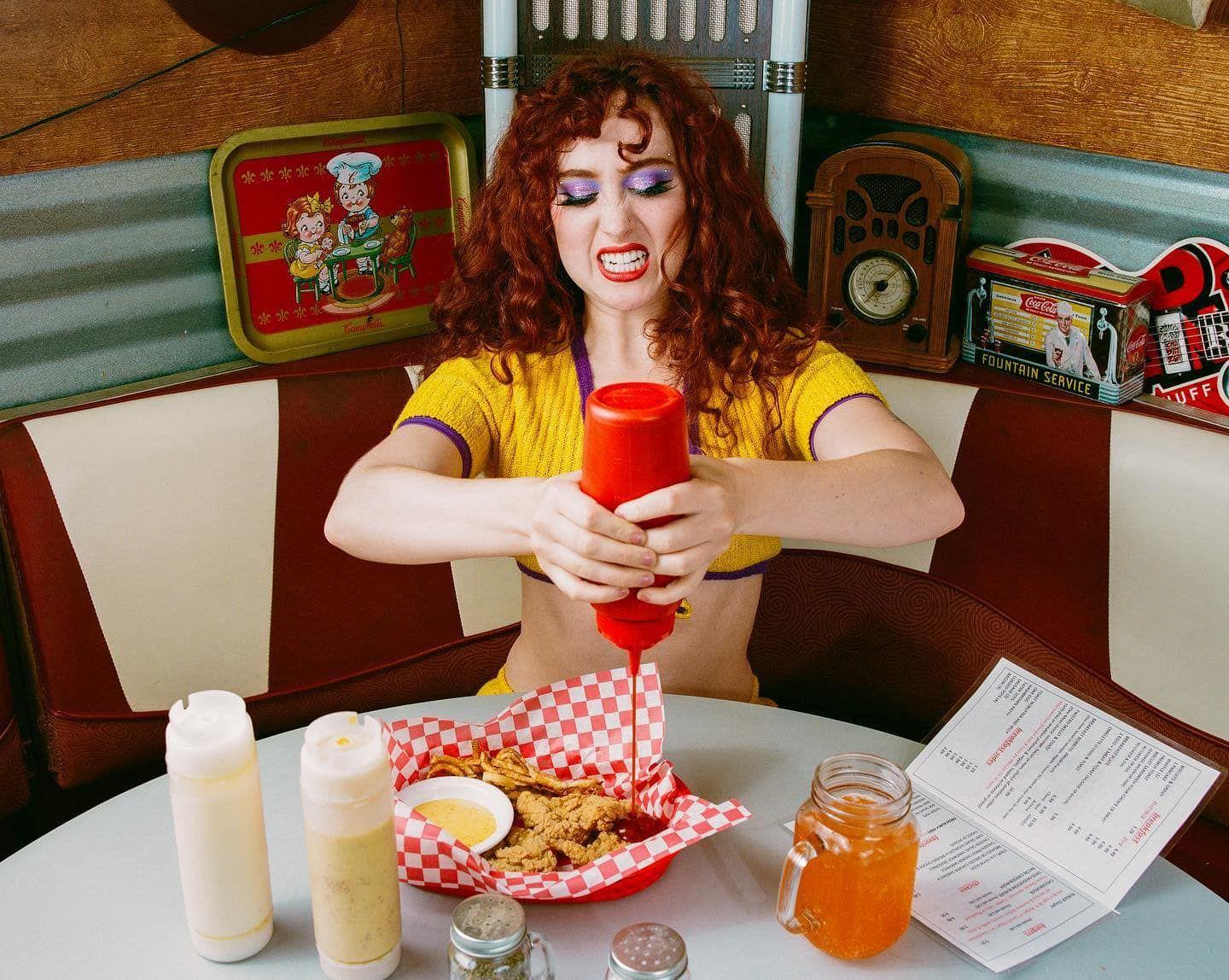 Chappell Roan em uma gravação de vídeo, sentado em uma lanchonete colocando ketchup no frango frito 