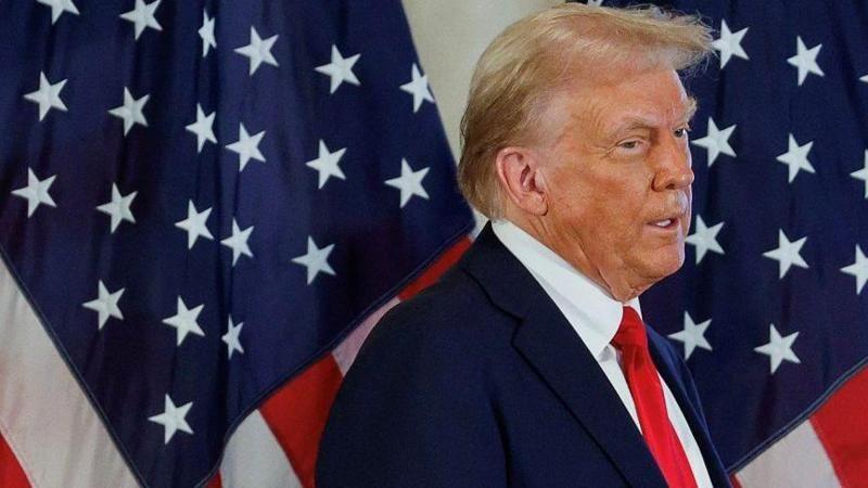 Trump de perfil em frente a bandeira dos EUA