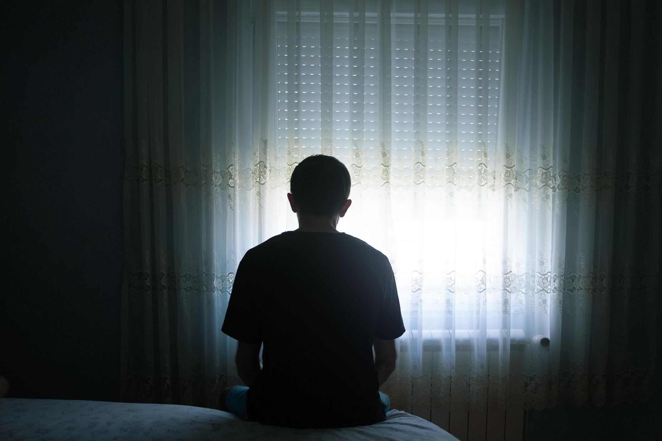 Un hombre deprimido en su cuarto a oscuras viendo la ventana