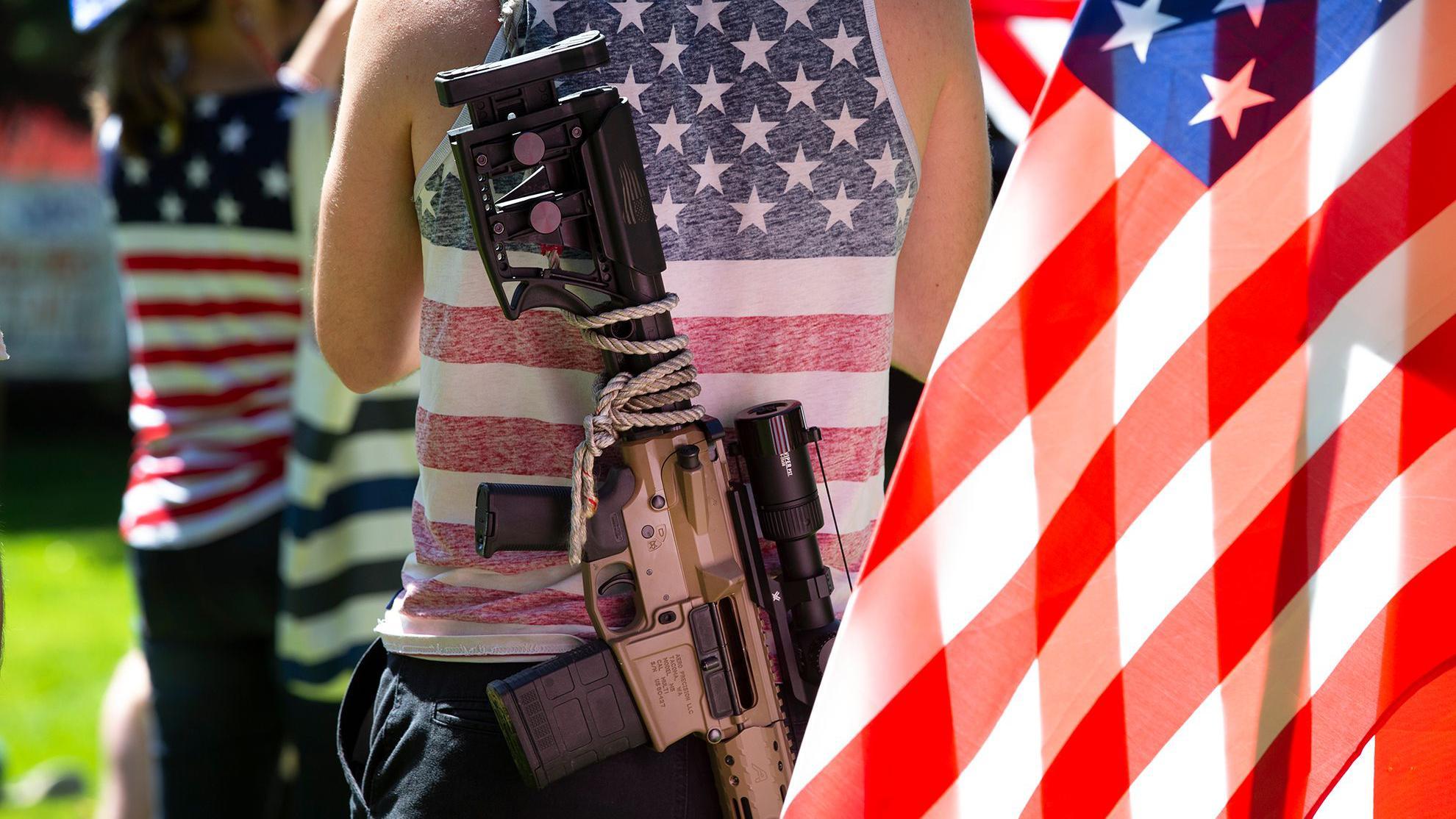 Hombre usa una camisa con la bandera de Estados Unidos y un rifle colgado en su espalda