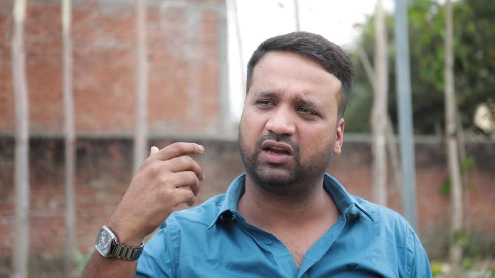 Zeeshan Ali gesticulando enquanto fala em área externa, perto dos muros de uma casa