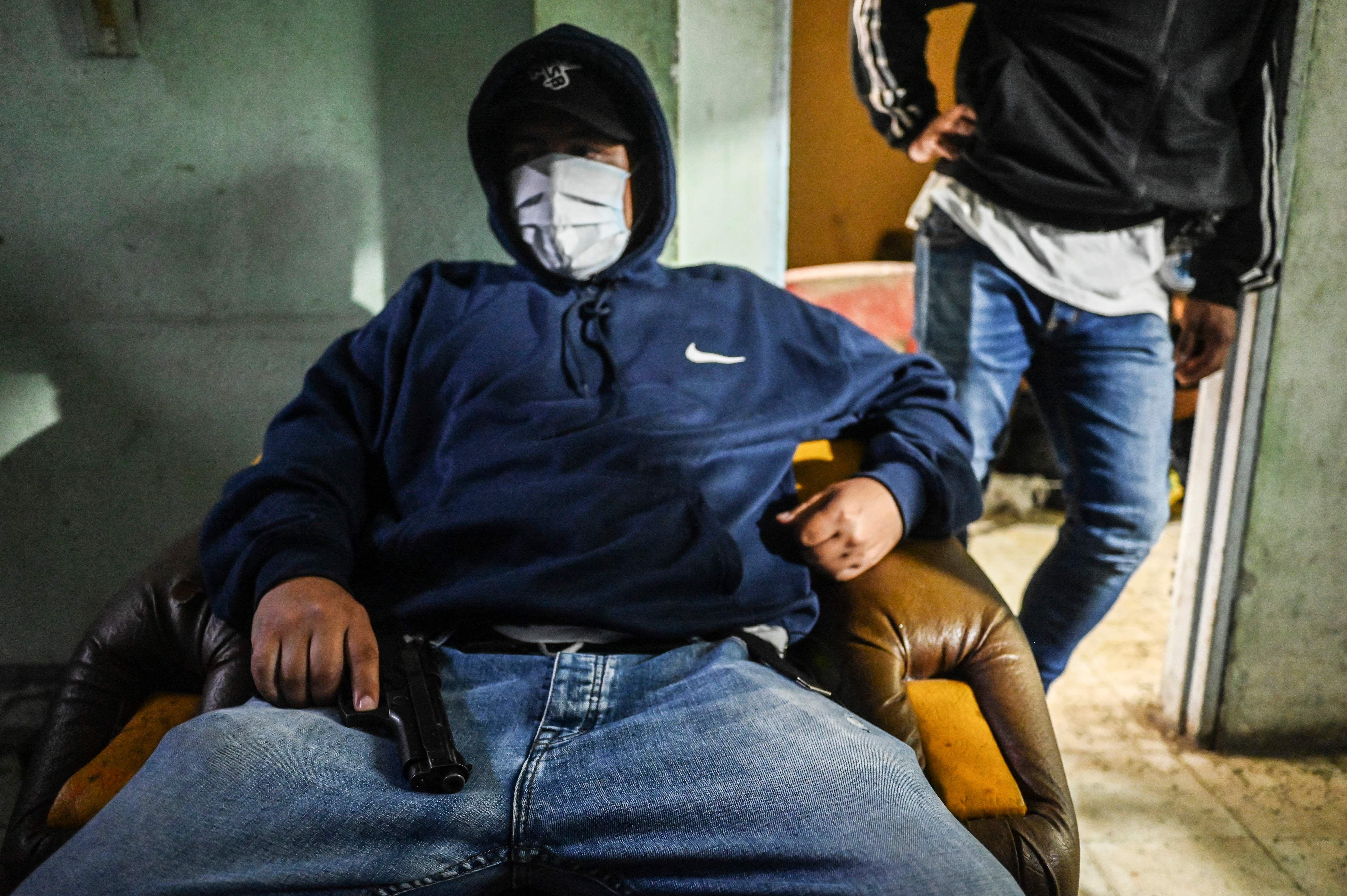 Un miembro de un grupo criminal en Colombia con el rostro cubierto sostiene un arma sentado en una silla.