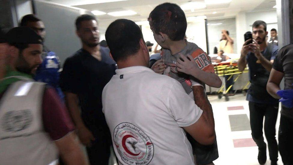 A Red Crescent aid worker carries a boy into hospital while people stand by and a man films the entry on his phone