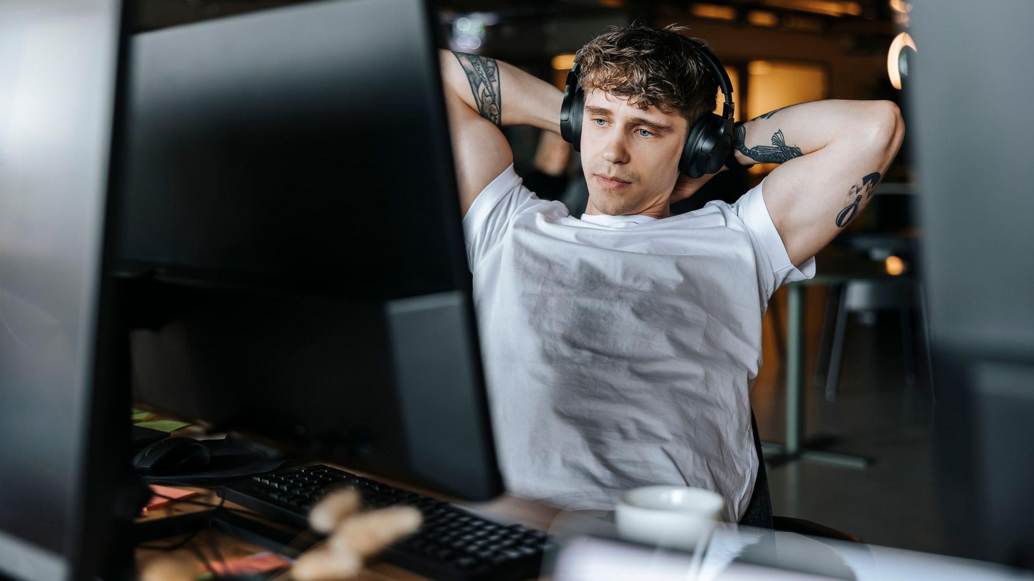 Hombre sentado frente a un escritorio con computadora, con los brazos hacia atrás en posición de relax y sus audífonos puestos.