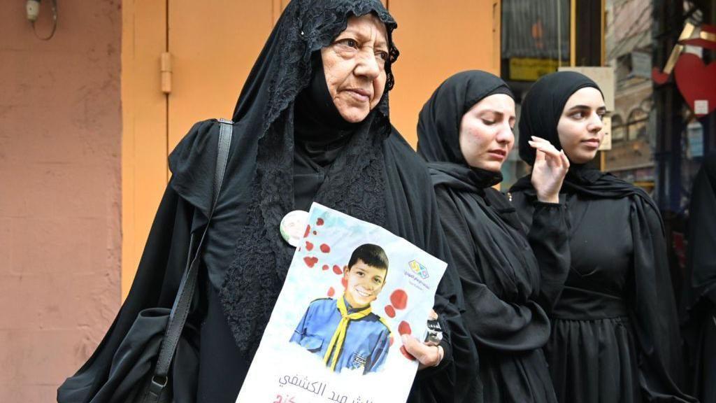 صورة لأم ترتدي الأسود بجوار أخريات، وتمسك بصورة أحد الطفلين اللذيْن قُتلا ضمن عشرات ضحايا هجمات أجهزة البيجر واللاسلكي المفاجئة.