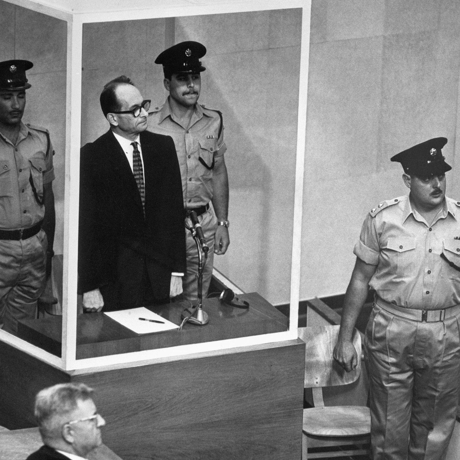 Adolf Eichmann se encuentra en su celda de cristal a prueba de balas para escuchar al Tribunal Supremo de Israel rechazar por unanimidad una apelación contra su sentencia de muerte. Junto a él hay dos guardias armados.