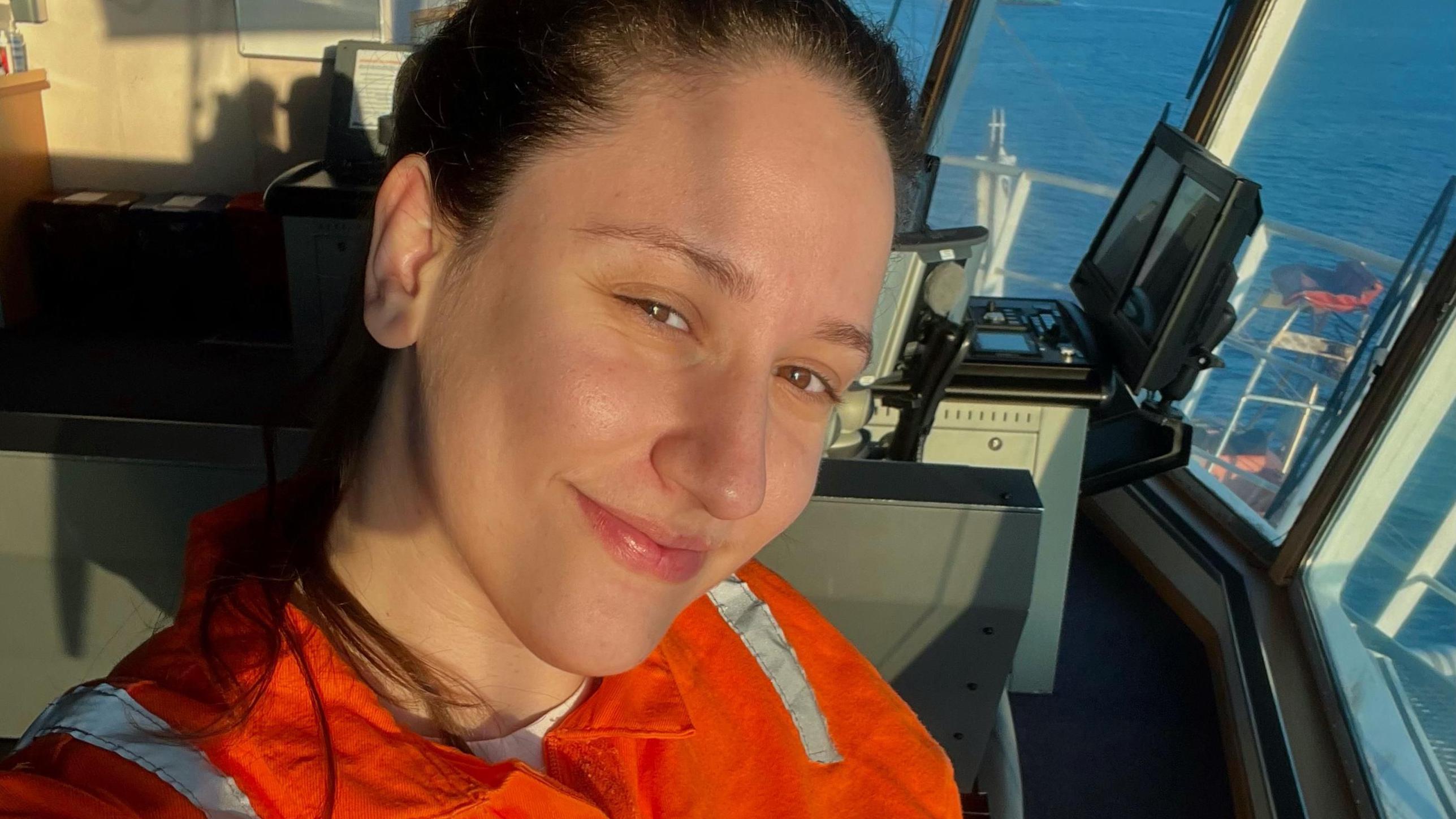 Karine com uniforme fazendo selfie sorrindo em frente a janela de navio, através da qual é possível ver o mar 