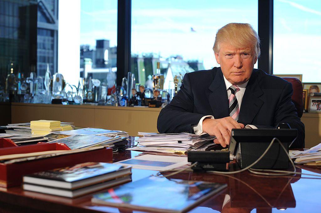 Donald Trump está sentado em sua mesa de escritório na Trump Tower, em Nova York. A mesa está coberta de revistas e jornais. O parapeito da janela atrás dele está repleto de prêmios, troféus e fotos emolduradas.