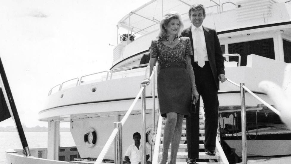 Foto em preto e branco de Donald e Ivana Trump saindo do iate Trump Princess. Ela está usando um vestido e ele está usando um terno escuro com uma gravata branca. O iate é branco e o mar está ao fundo.