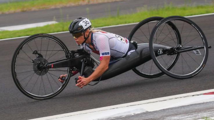 A foto mostra uma mulher de pele clara, com capacete preto em uma cadeira adaptada para o ciclismo paralímpico. Ela pedala com os braços.