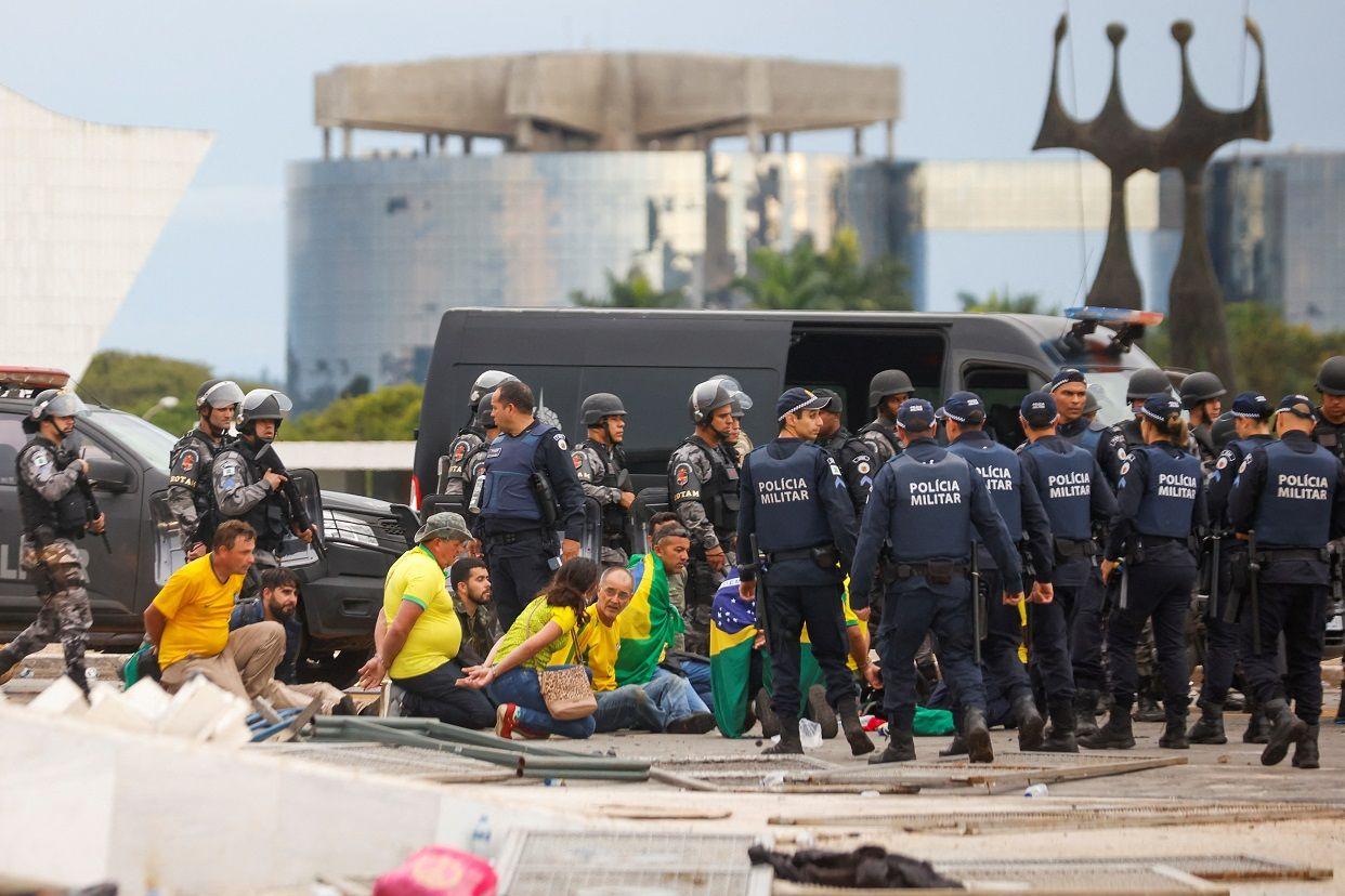 Pessoas com camisas verdes e amarelas sentadas com as mãos algemadas; eles são cercados por policiais na Praça dos Três Poderes