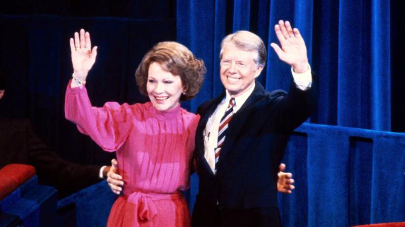 Jimmy Carter y Rosalynn Carter saludan a la multitud, mientras con la otra mano cada uno agarra al otro por la cintura. 