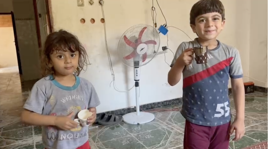 A girl standing on the left and a boy standing on the right sip tea at home in Gaza.