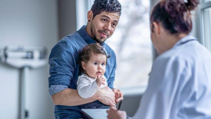 Homem com bebê no colo, observado por uma médica em consultório