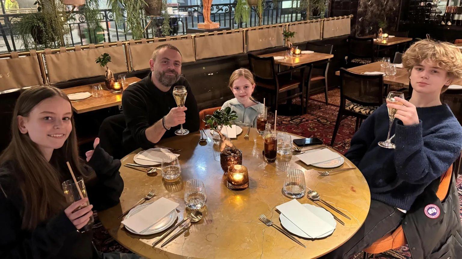 Se ve a Geraint sentado con sus tres hijos en un bonito restaurante: a uno de sus lados, la niña mayor mira la cámara sonriendo, luego está Gerain, después su hija menor, quien está sentada sonriendo, y luego su hijo mayor, quien está serio con el vaso en alto.