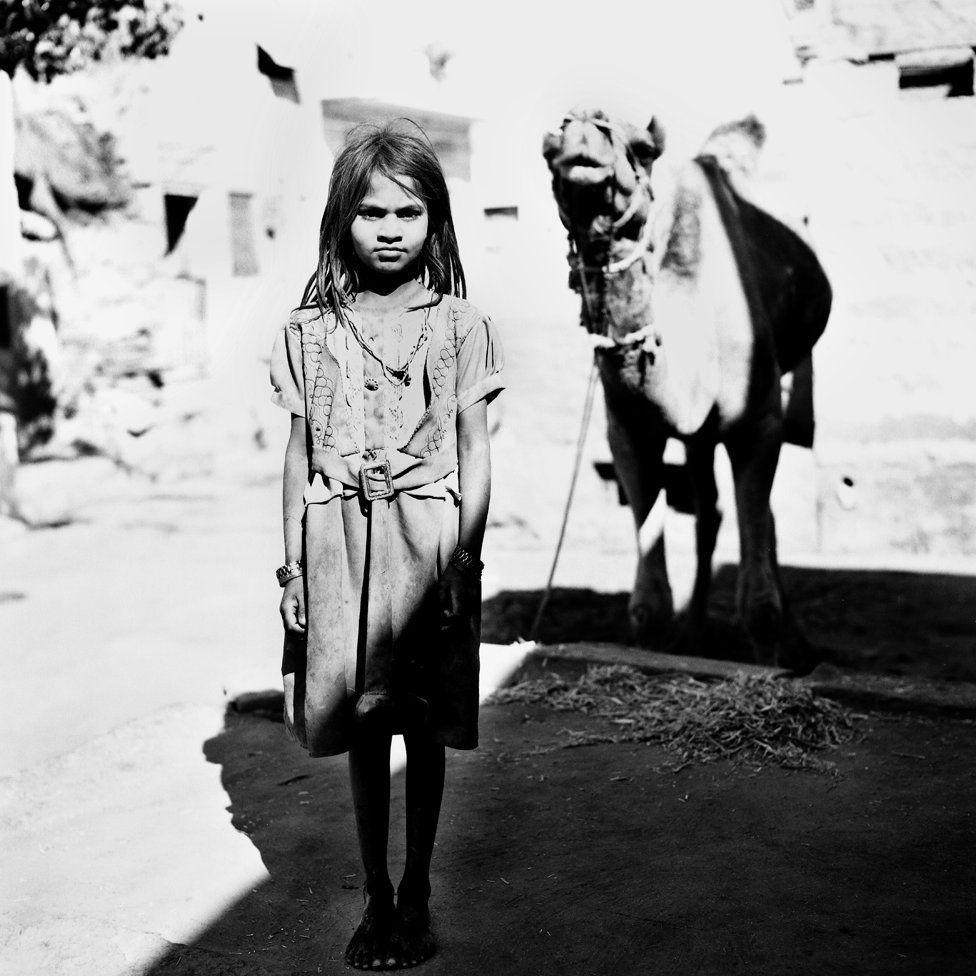 Garota posa para foto, com um camelo ao fundo