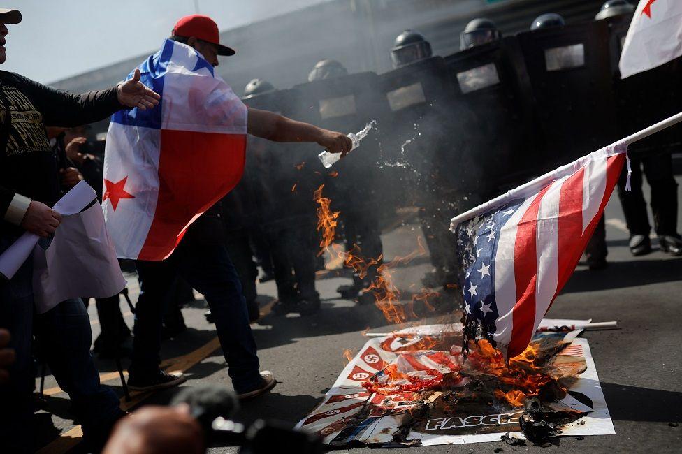 Um homem com a bandeira do Panamá nas costas joga água em uma bandeira de chama.