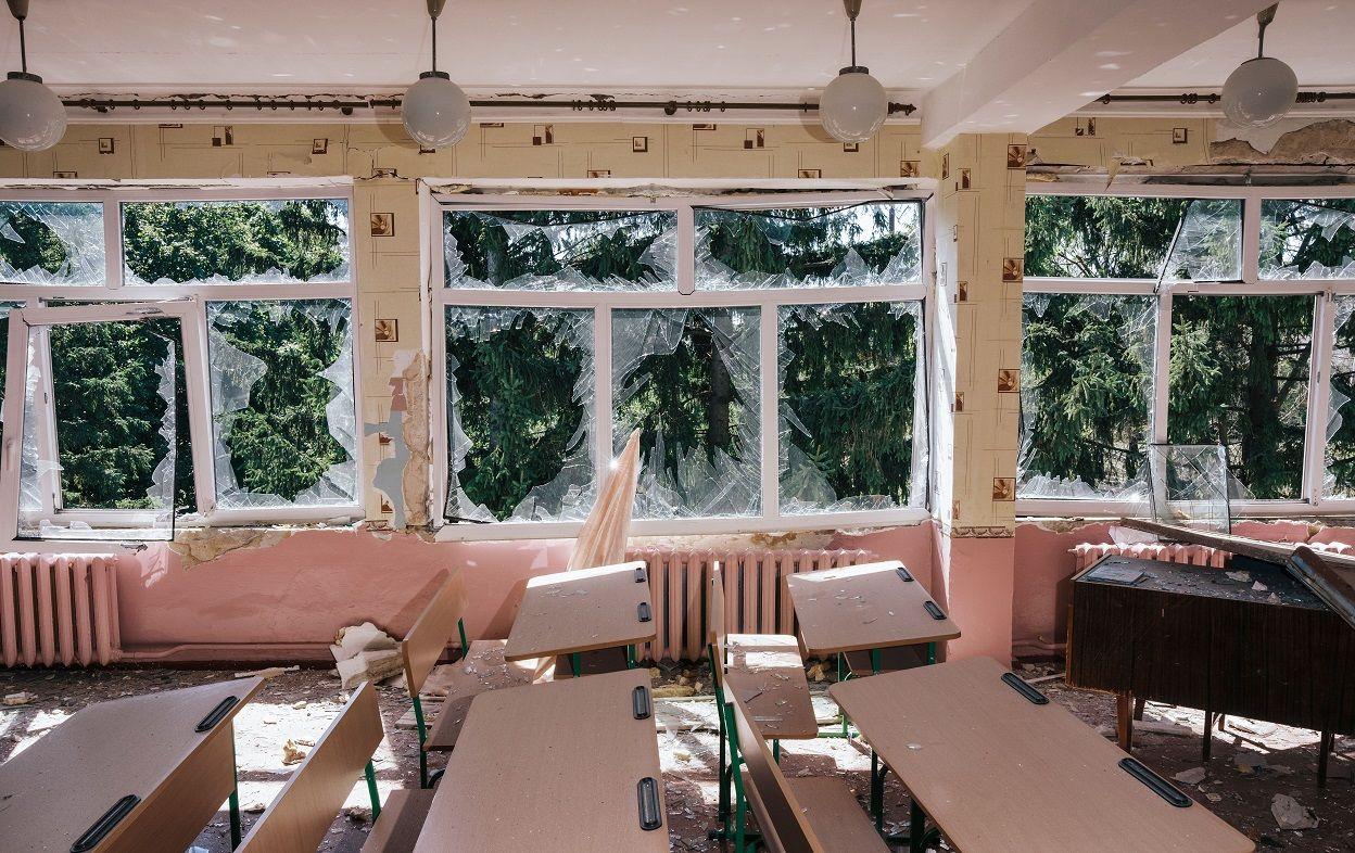 A foto mostra uma sala de aula, com carteiras e bancos, e três grandes janelas totalmente destruídas. 