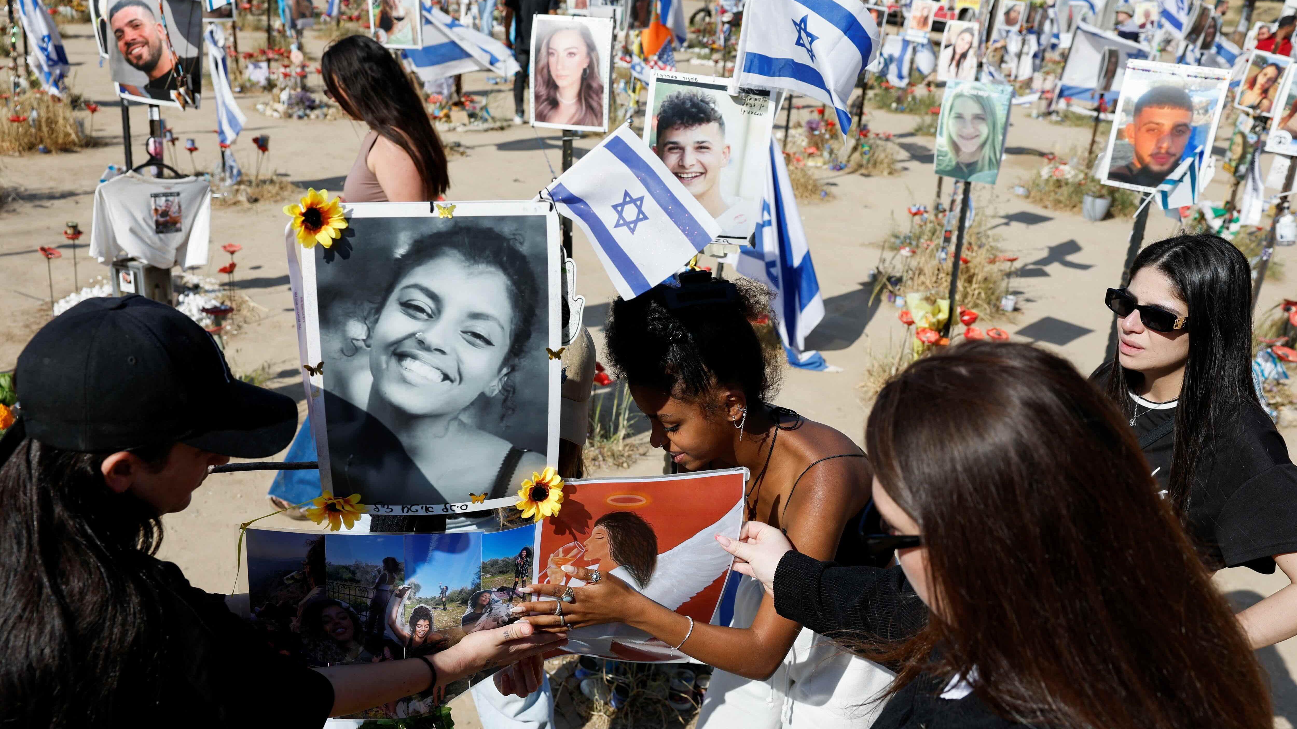 Pessoas agachadas prestam homenagem às vítimas com fotos e bandeiras israelenses em área aberta, com aparência de deserto