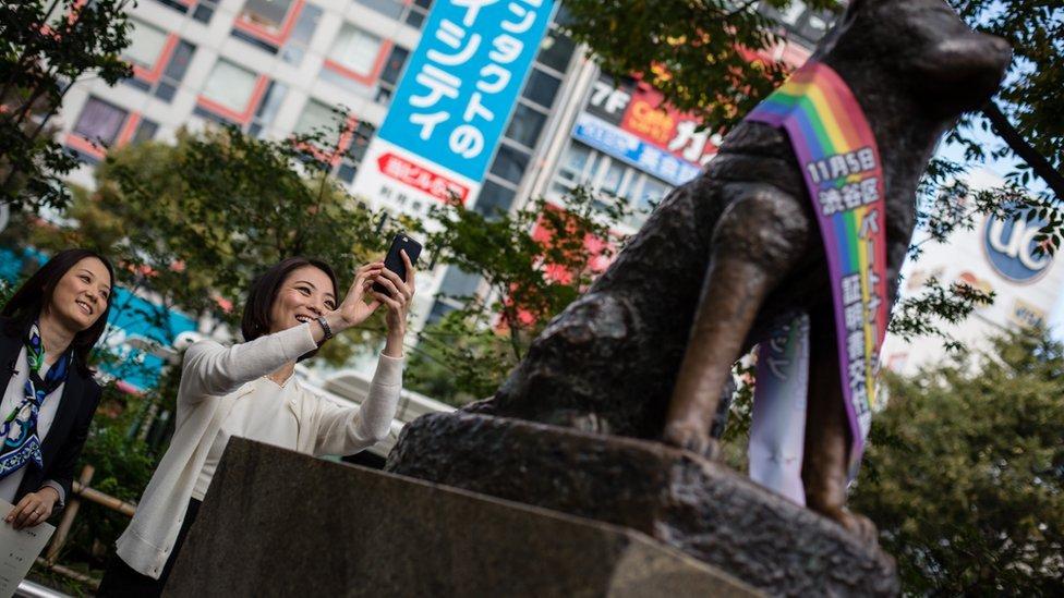 Mulher tirando foto da estátua de Hachiko que está com uma faixa com as cores do arco-íris