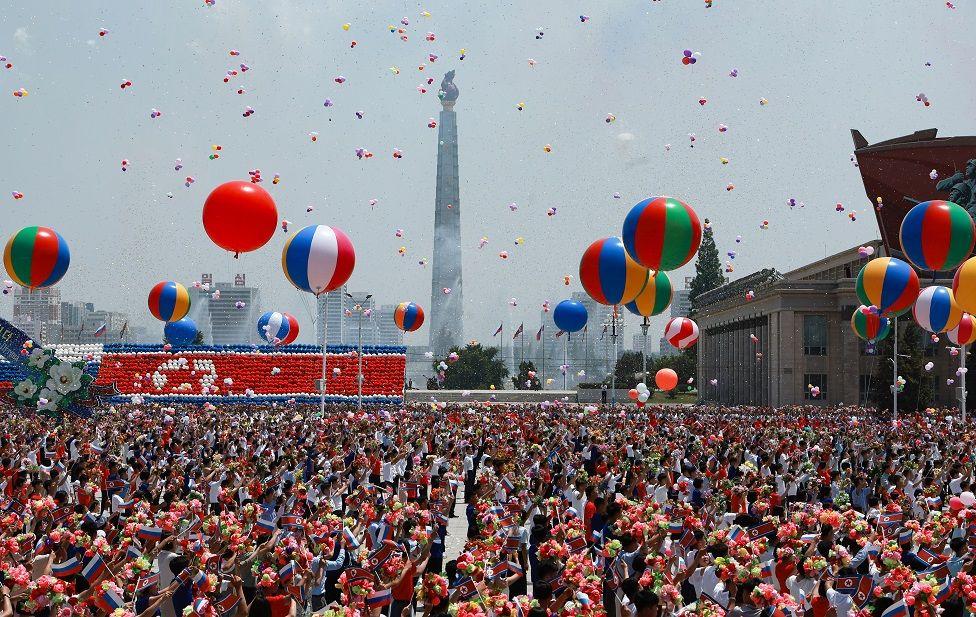 Una multitud con globos de colores y flores en una amplia plaza 