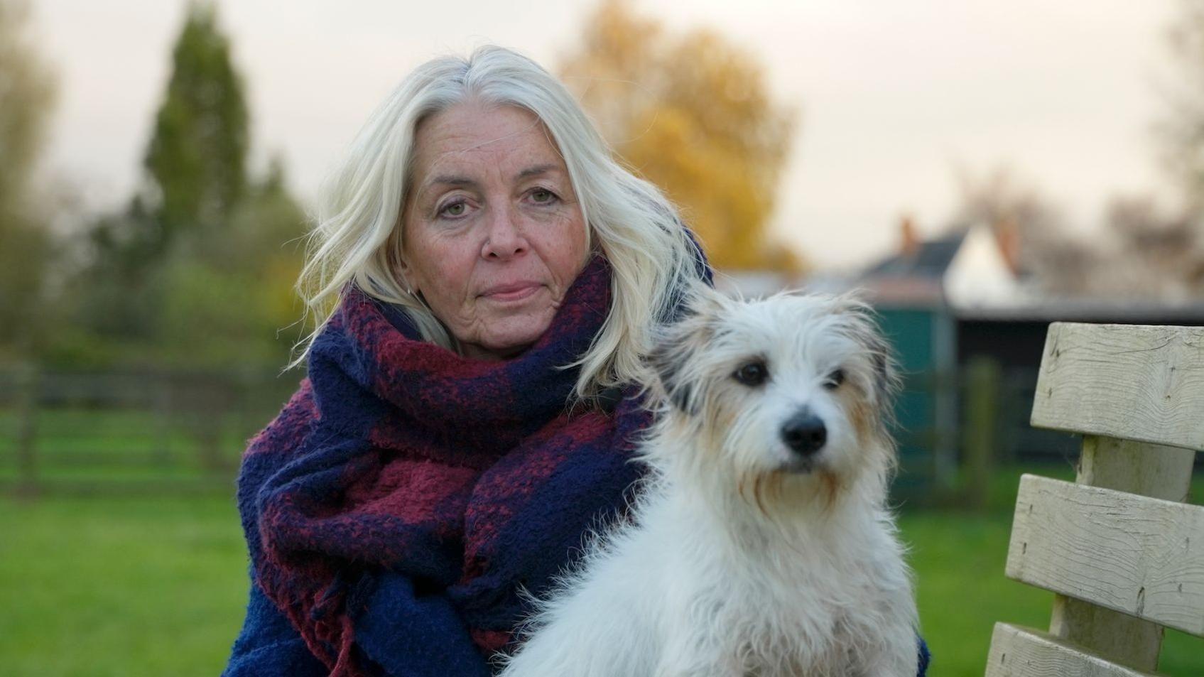 Lisa Willis, una mujer de pelo rubio claro, envuelta en una gran bufanda azul y violeta y un abrigo azul intenso, está sentada en un banco al aire libre, con césped, árboles y un puñado de tejados visibles detrás de ella. Duke, su terrier, que tiene el pelo blanco con manchas grises y marrones alrededor de las orejas, los ojos y el hocico, está sentado a su lado.