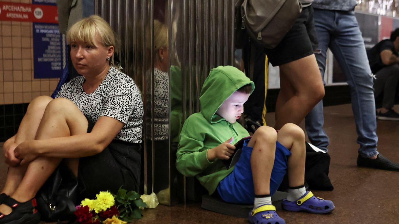 Un niño con una tableta, junto a una mujer y otras personas refugiadas en el metro de Kyiv. 