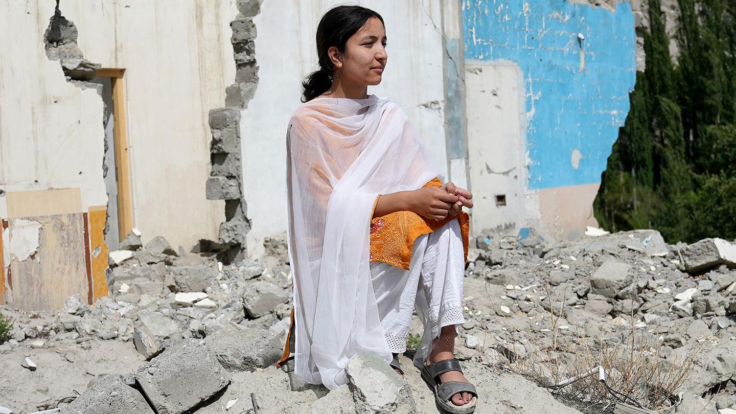 Komal Sher looks out to the right of the camera while wearing an orange top, snadals and white shawl. She is standing on the rubble of buildings in her village of Hassanabad, with damaged walls in the background.