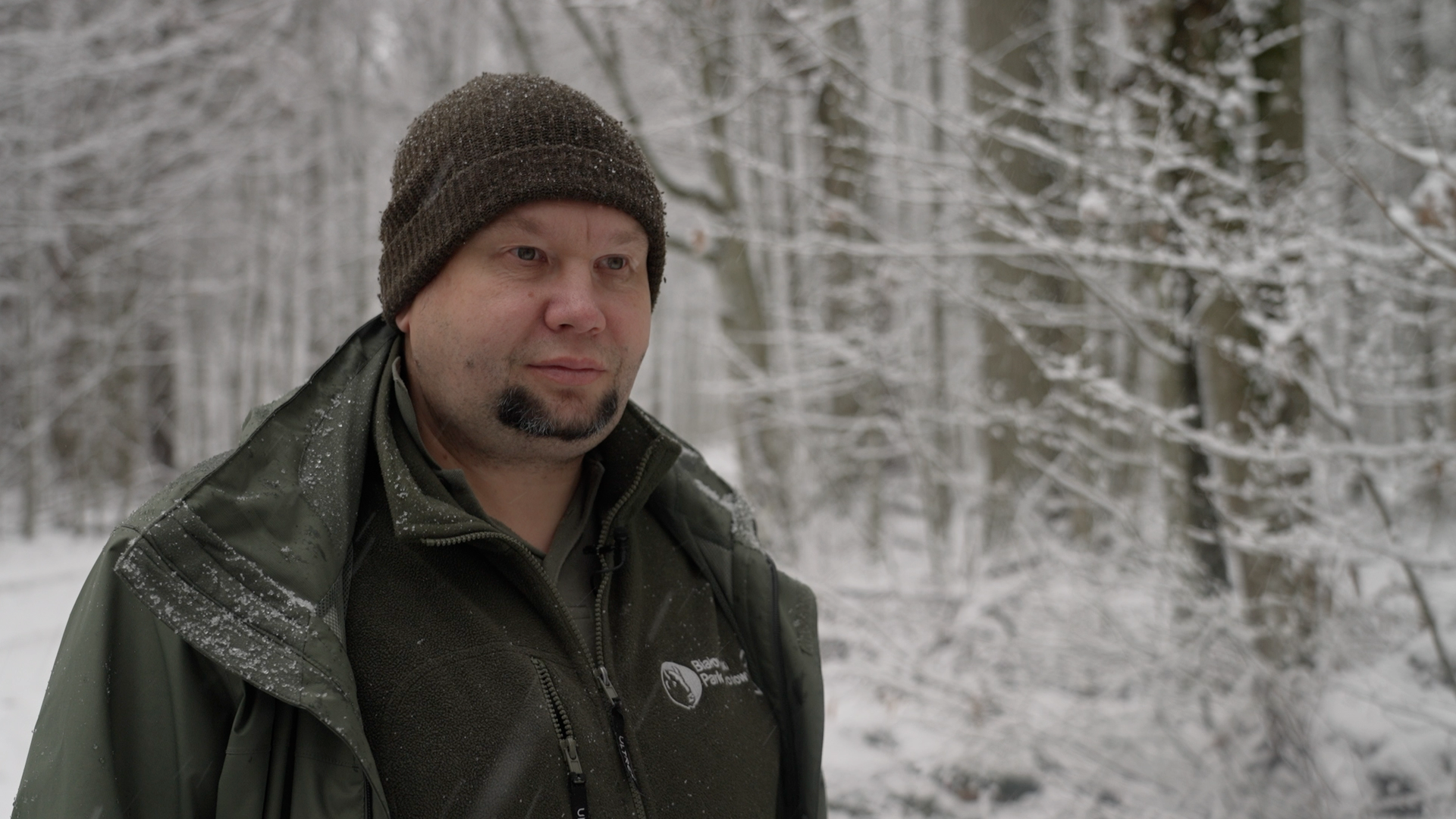 A man stands in the forefront of the image wearing a beanie, jacket and fleece jumper - all are dark green in colour.