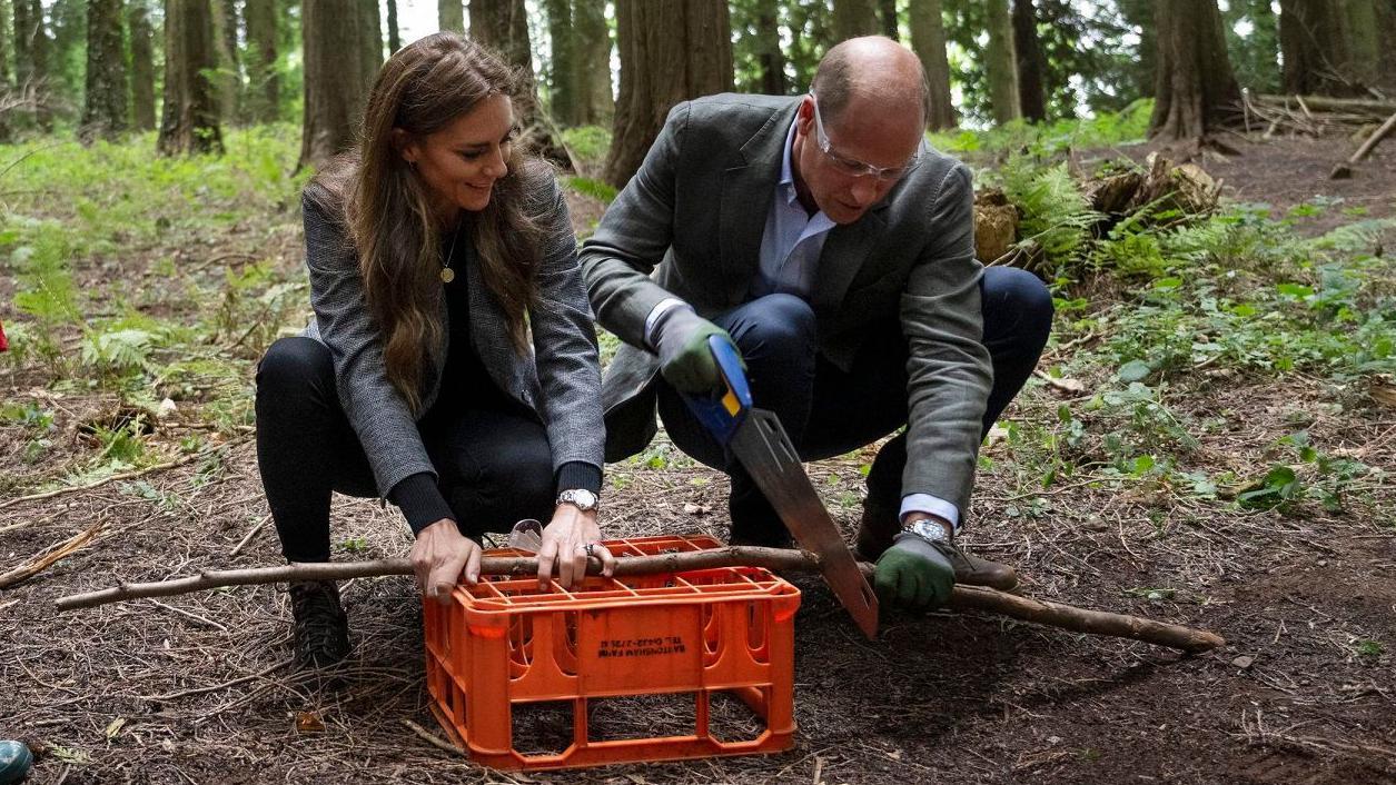 Kate e William agachados em meio à floresta; ele corta pedaço de lenha, e ela ajuda