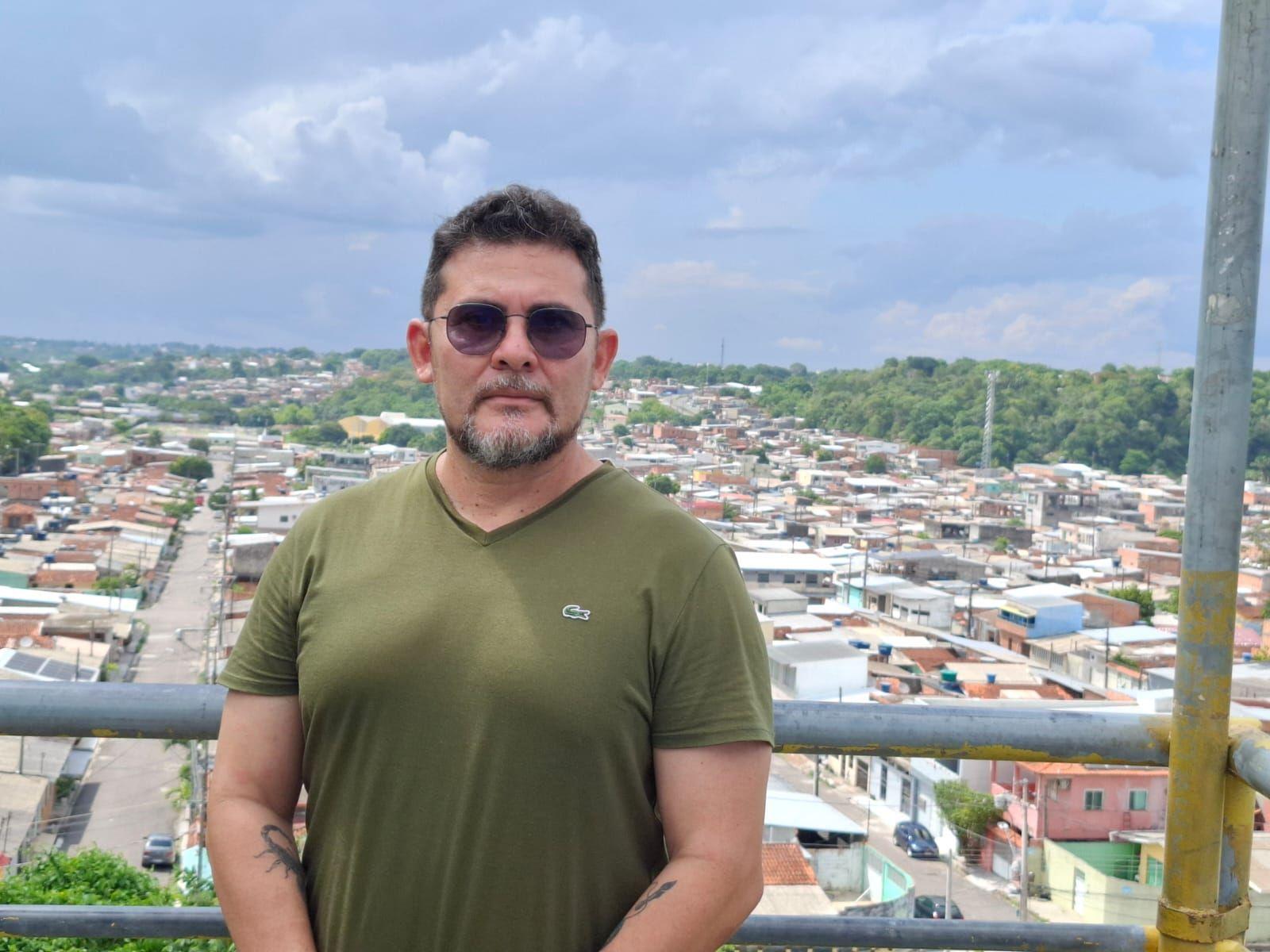 Profº Marcos Castro na Cidade de Deus posa para foto em local elevado na Cidade de Deus, com casas visíveis abaixo, em segundo plano