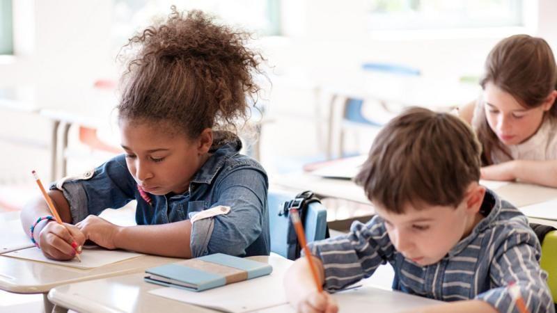 Crianças multiétnicas escrevem em cadernos em suas carteiras escolares
