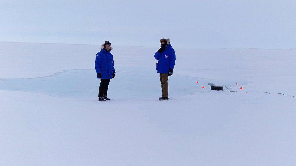 Dos investigadores envueltos en equipo ártico mientras observan la bomba de agua de mar en acción.