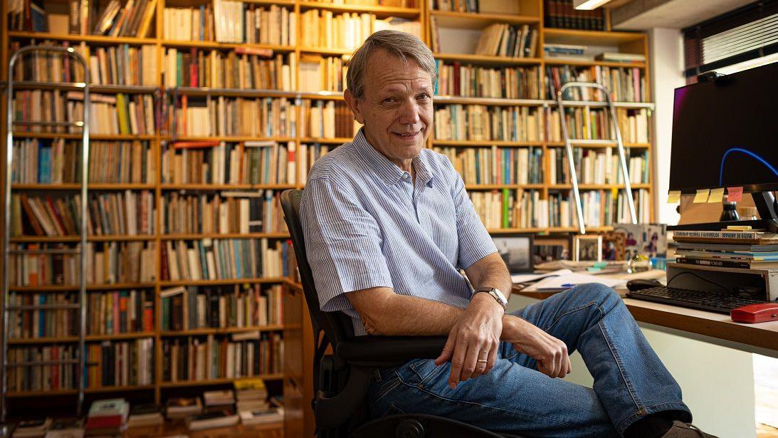 Foto de André Singer sentado em uma cadeira com uma grande prateleira de livros atrás dele. 