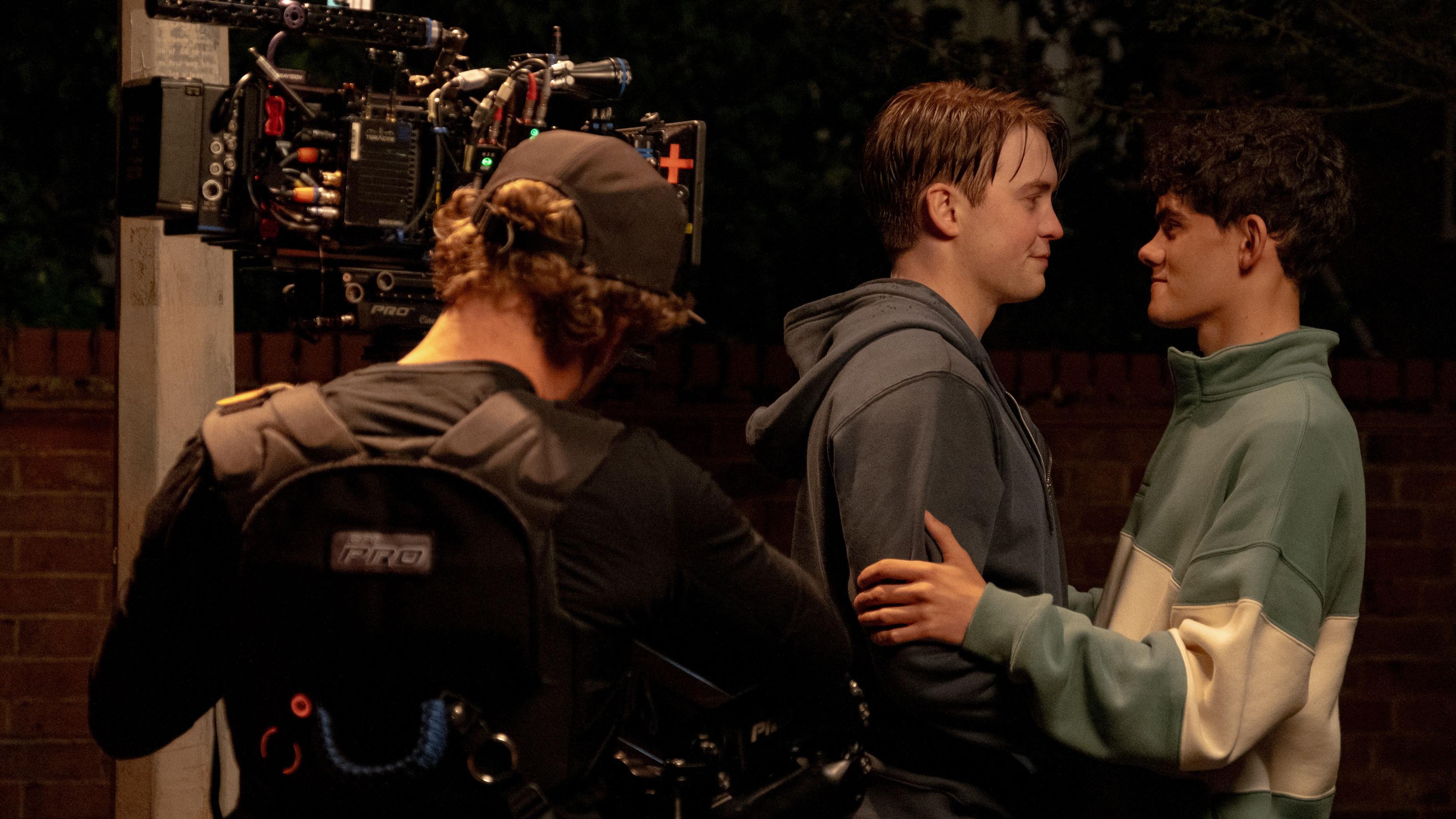 Um operador de câmera, usando um boné e de costas para o fotógrafo, filma Kit Connor e Joe Locke enquanto se abraçam e sorriem um para o outro. O cabelo ruivo de Kit está molhado e ele veste um moletom cinza, enquanto Charlie tem cabelos castanho-escuros e usa um suéter largo verde e branco.