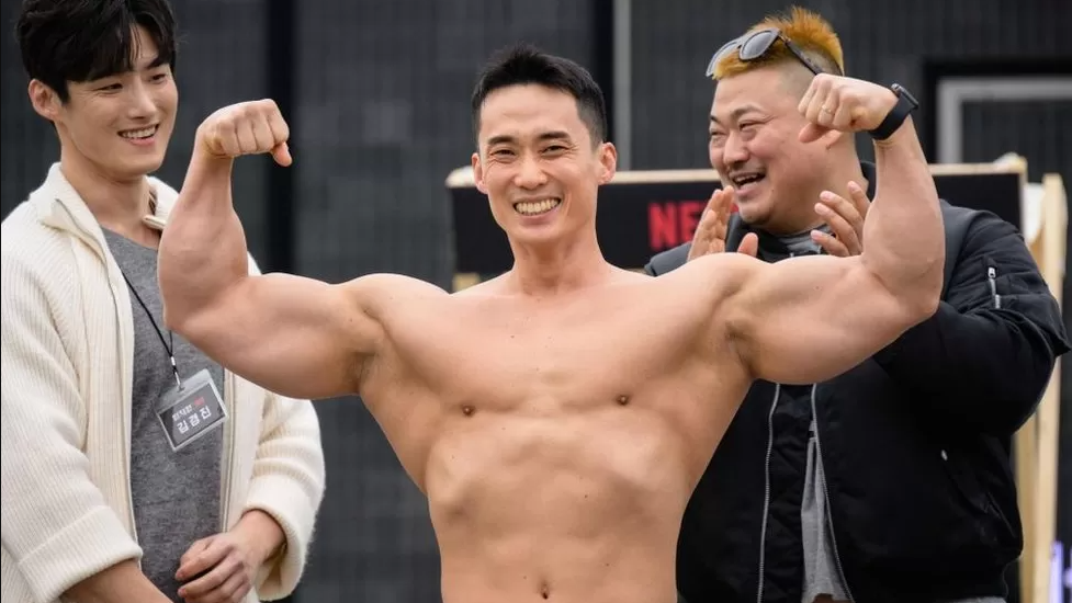 Homem erguendo os músculos e sorrindo, com outros 2 homens atrás sorrindo