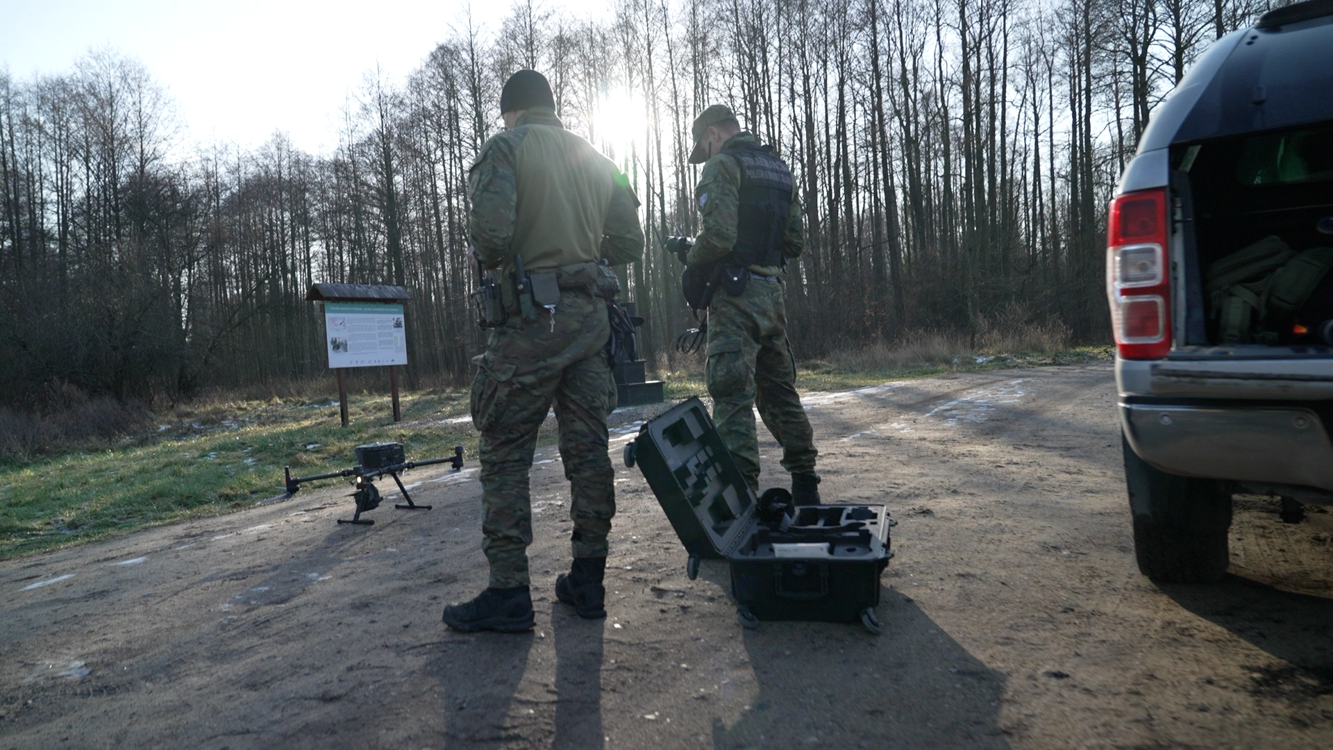 Two men dress in camouflage look done at a drone which sits on the floor in a clearing in the woodland.