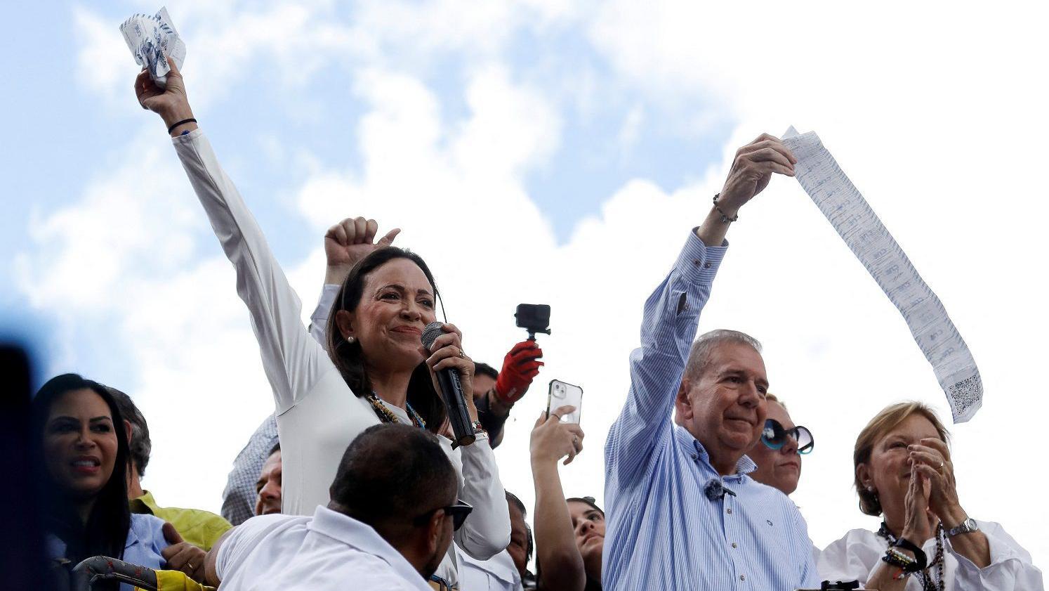 María Corina Machado e Edmundo Gonzalez levantam os braços, rodeados de outras pessoas, em cima de um carro de som