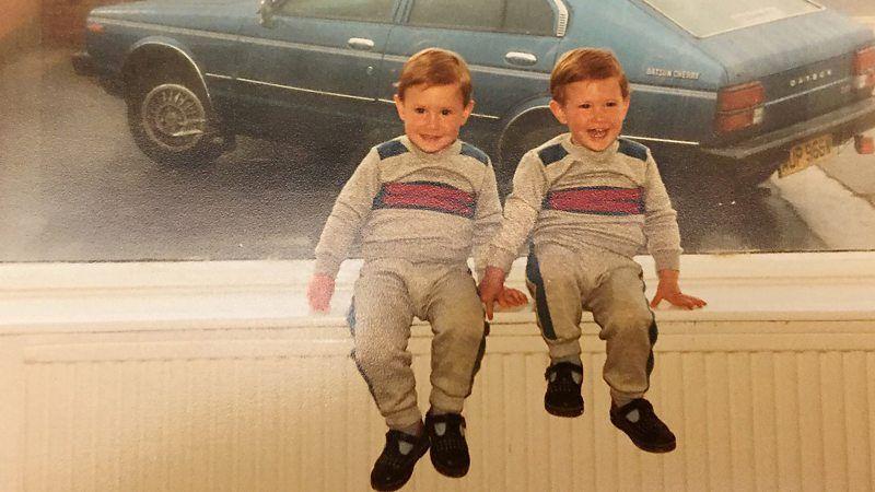 Fotografia antiga mostra dois meninos brancos, gêmeos idênticos, loiros, usando a mesma roupa, sentados em uma plataforma e sorrindo 