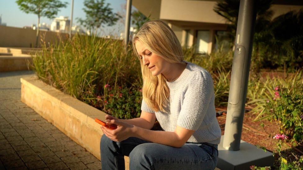 Kate Konlin sentada em mureta ao ar livre usando o celular