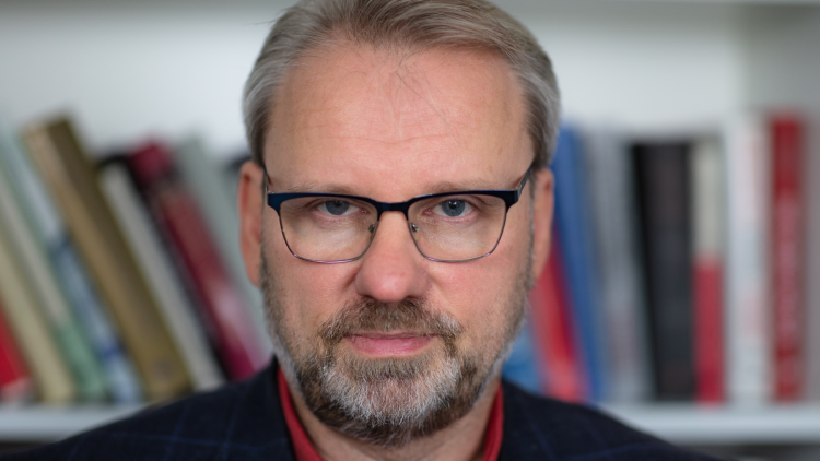 Wolfgang Münchau con gafas y barba delante de una librería llena de libros. 