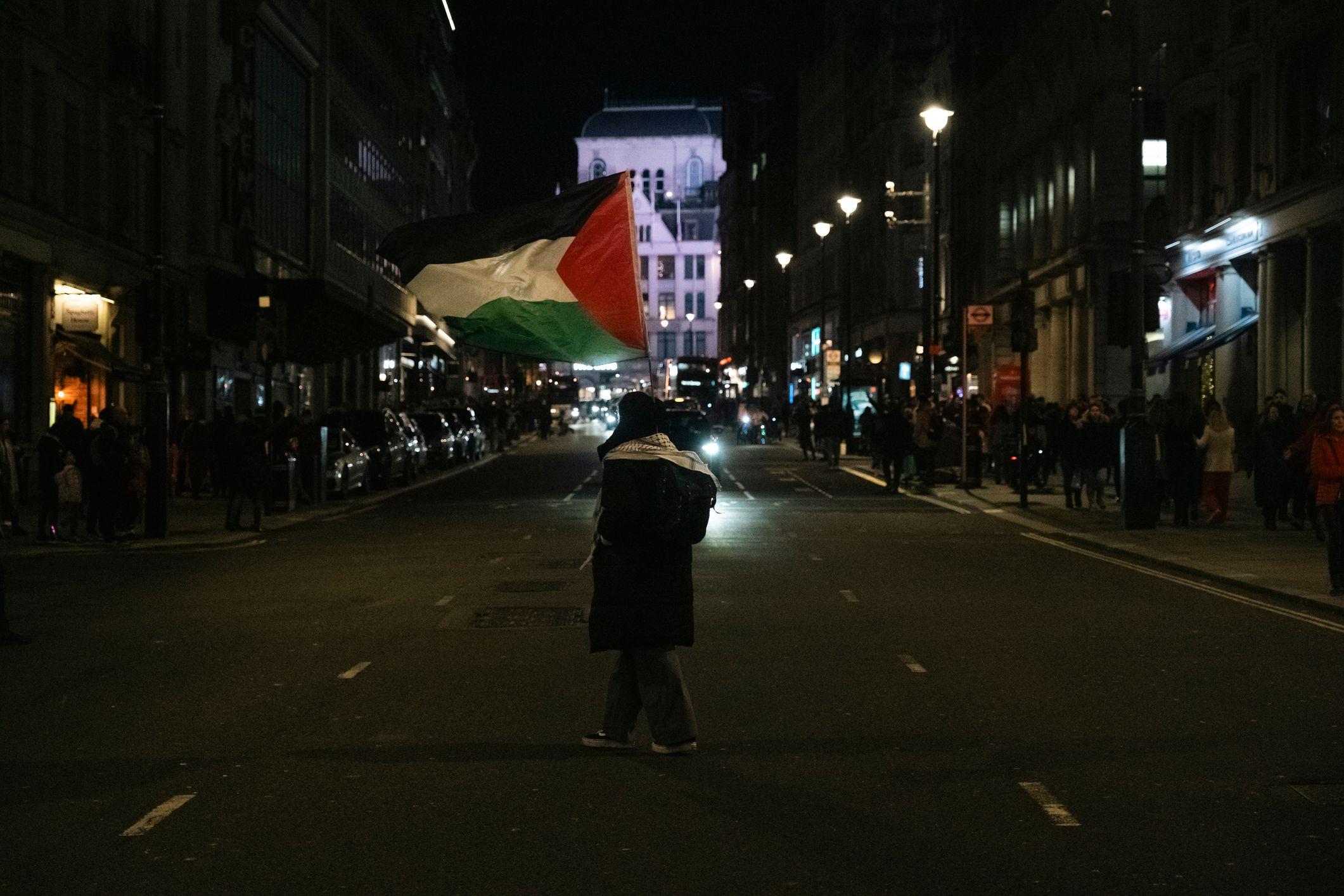 شابة تمشي في شوارع لندن، تحمل علم فلسطين وترتدي الكوفية الفلسطينية. الصورة التقطت ليلاً ولا تظهر فيها ملامح الفتاة.