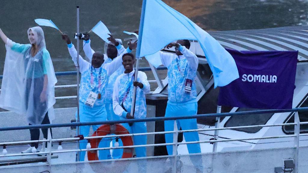 Ali Idow Hassan agitando uma bandeira em um barco com membros da delegação olímpica da Somália