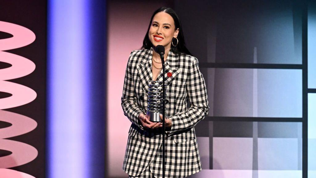 Meena Harris vestida com um terno xadrez preto e branco, falando ao microfone ao receber uma premiação em evento em Nova York