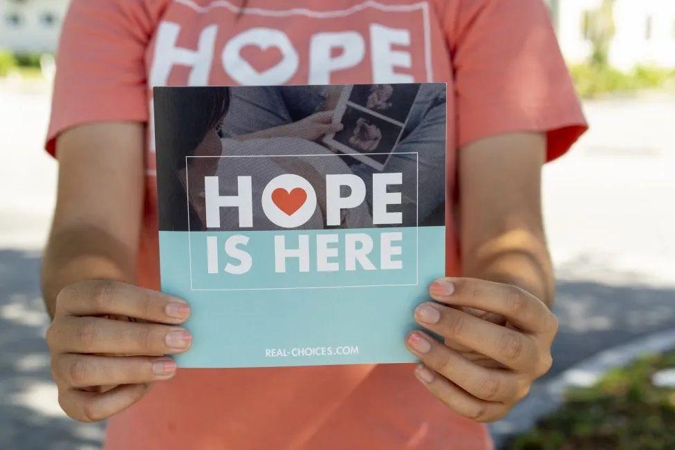Mão de mulher segurando cartaz em que se lê: 'A Esperança está aqui', em inglês