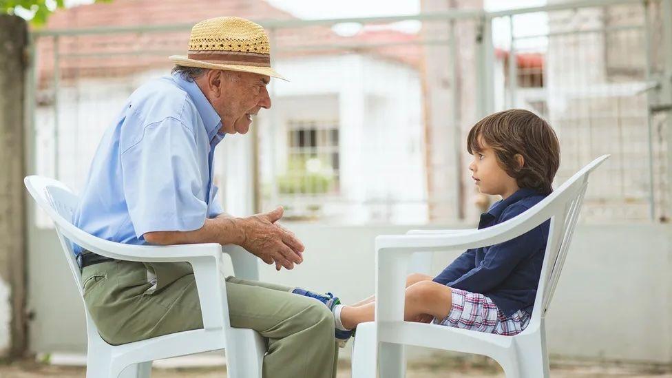Un hombre mayor está sentado hablando con un pequeño niño que lo escucha con atención