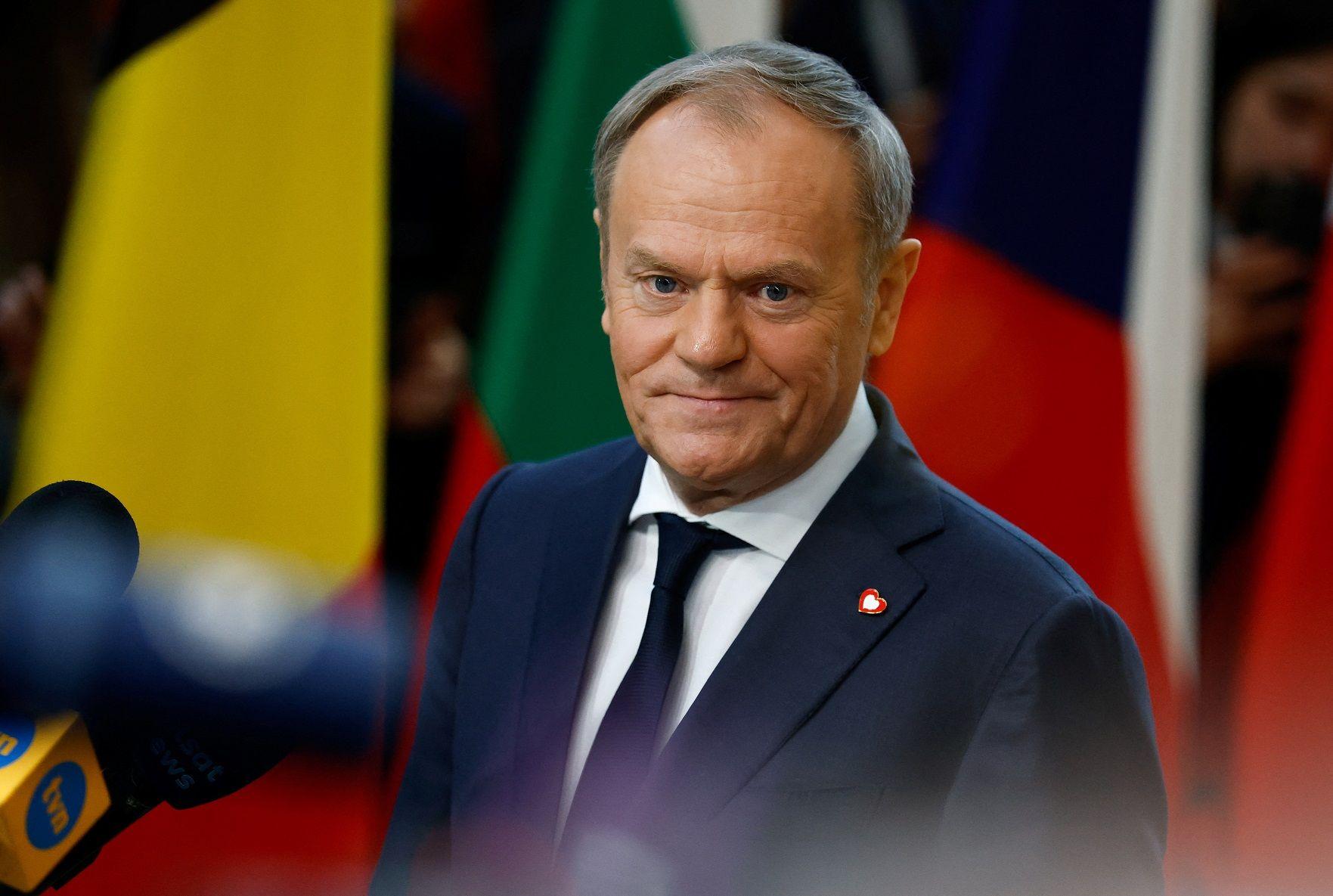 Donald Tusk em pé, rodeado por microfones da imprensa, com olhar compenetrado