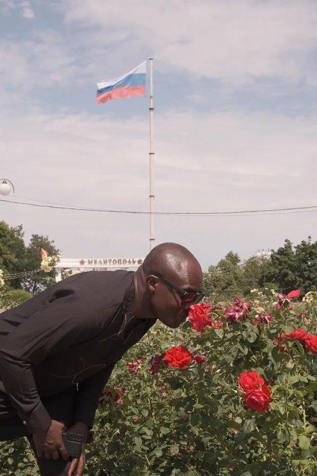 Jornalista fotografado cheirando a flor de uma roseira, com a bandeira russa tremulando ao fundo.