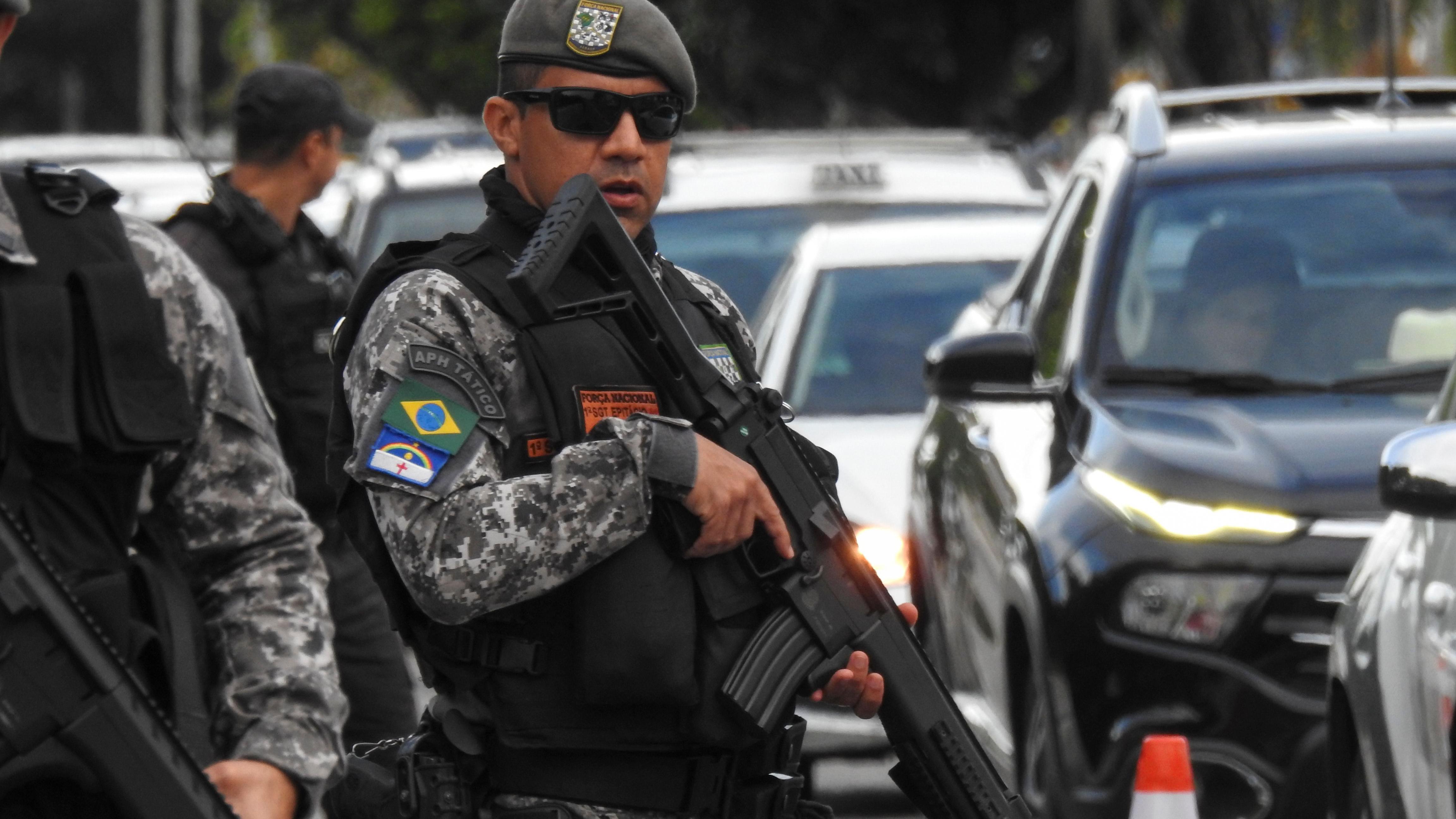 Policial usando óculos escuros segura arma de grosso calibre próximo a carros de polícia