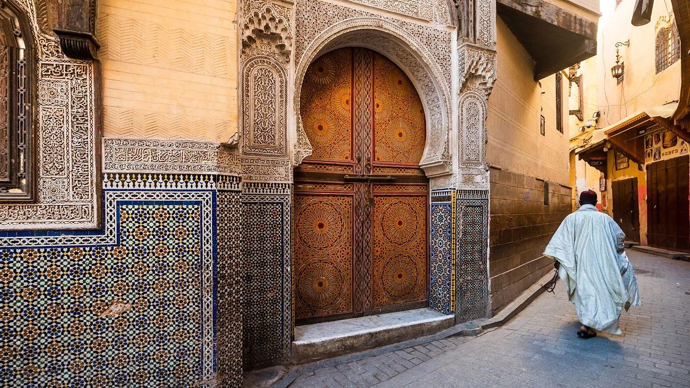 Um homem caminhando por um caminho estreito com uma grande porta de madeira e paredes de azulejos. 